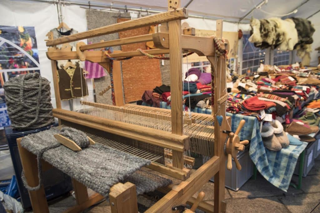 Ein traditioneller Webstuhl aus Holz mit grauem Gewebe im Vordergrund, umgeben von Wolle und bunten Textilien auf einem Marktstand in einem Zelt.