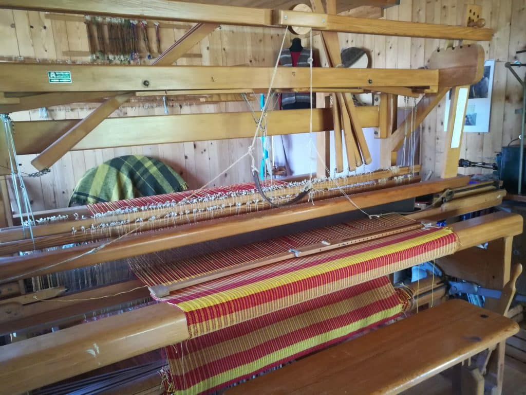 Ein traditioneller Webstuhl mit einem halbgewebten roten und gelben Stoff, umgeben von Holzwänden.