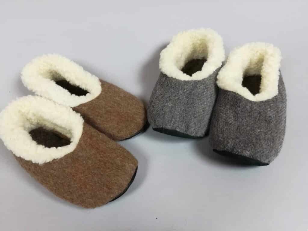 Zwei Paar Hausschuhe mit Filzoberfläche, eines in Braun und eines in Grau, beide mit weißem, flauschigem Innenfutter, auf einem weißen Hintergrund.