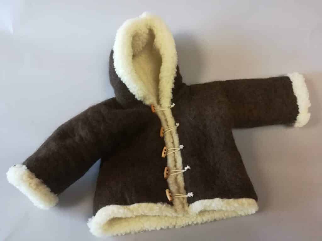 Kuschelige braune Jacke aus Wolle mit Kapuze, weißen Plüschbesätzen und Knebelknöpfen auf einer flachen Oberfläche liegend.