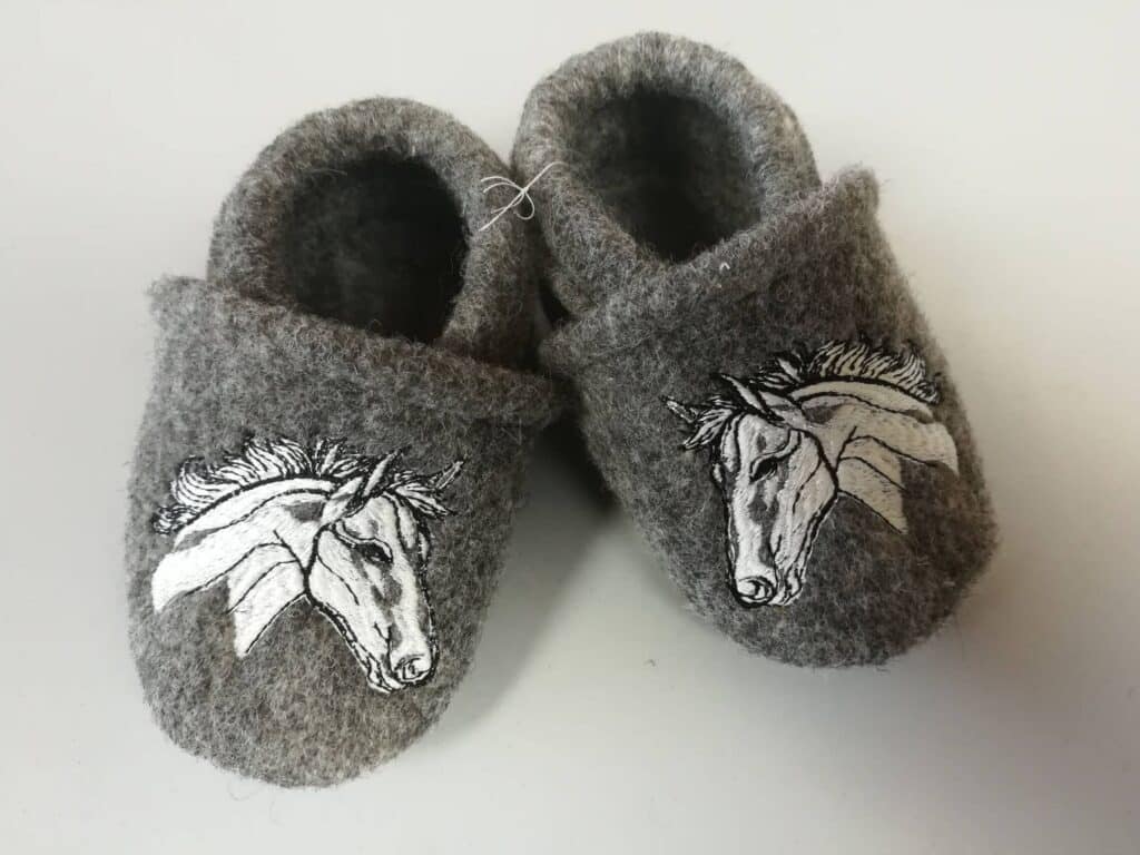 Graue Filzpantoffeln mit einem weiß-schwarz gestickten Pferdekopf-Design auf der Oberseite, auf einem weißen Hintergrund.
