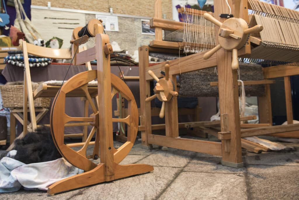 Ein traditionelles Spinnrad und Webstuhl aus Holz in einer Werkstatt, umgeben von Garnen und Textilien.