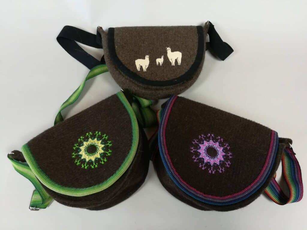 Drei braune Umhängetaschen aus Filz mit kettenartigen Handtaschendesigns. Eine hat weiße Tiermotive, die anderen zwei haben bunte, kreisförmige Muster mit grünen und lila Details sowie grünen und gestreiften Riemen.