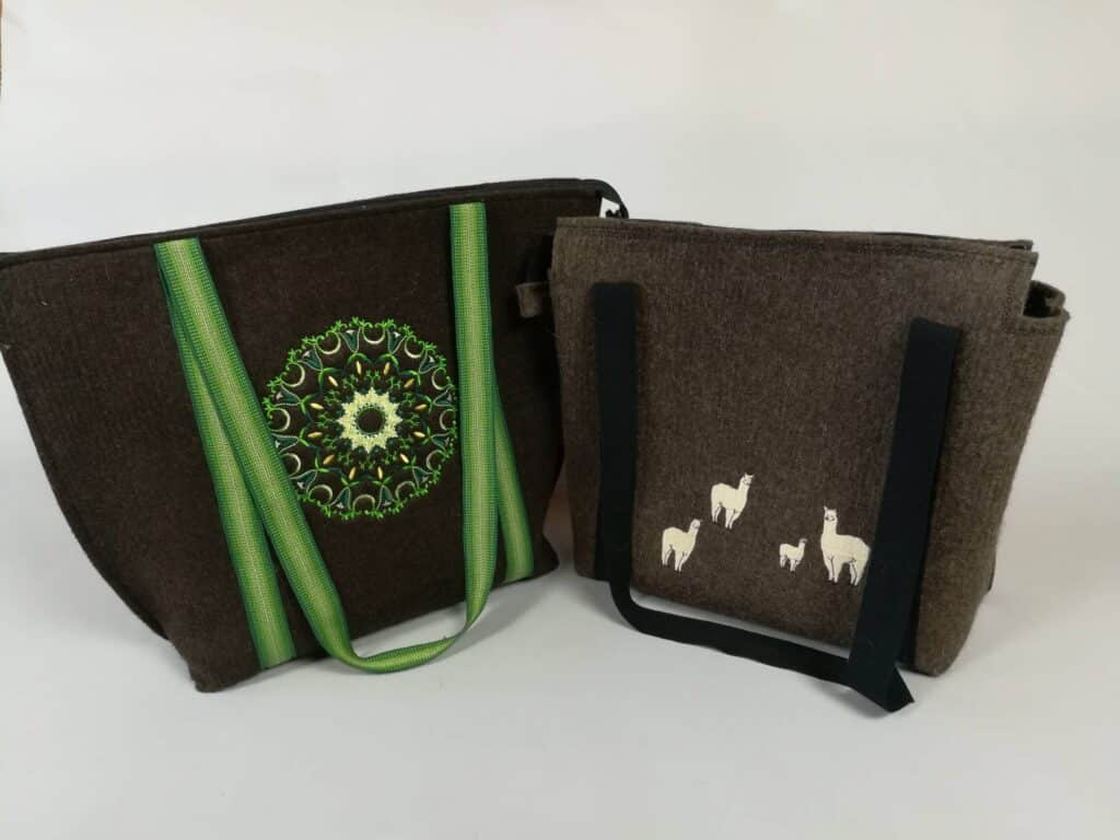 Zwei Filztaschen nebeneinander. Die linke Tasche ist dunkelbraun und hat grüne Tragegriffe sowie ein grünes, kreisförmiges Stickmuster. Die rechte Tasche ist ebenfalls dunkelbraun, hat schwarze Tragegriffe und ist mit weißen Lamas verziert.