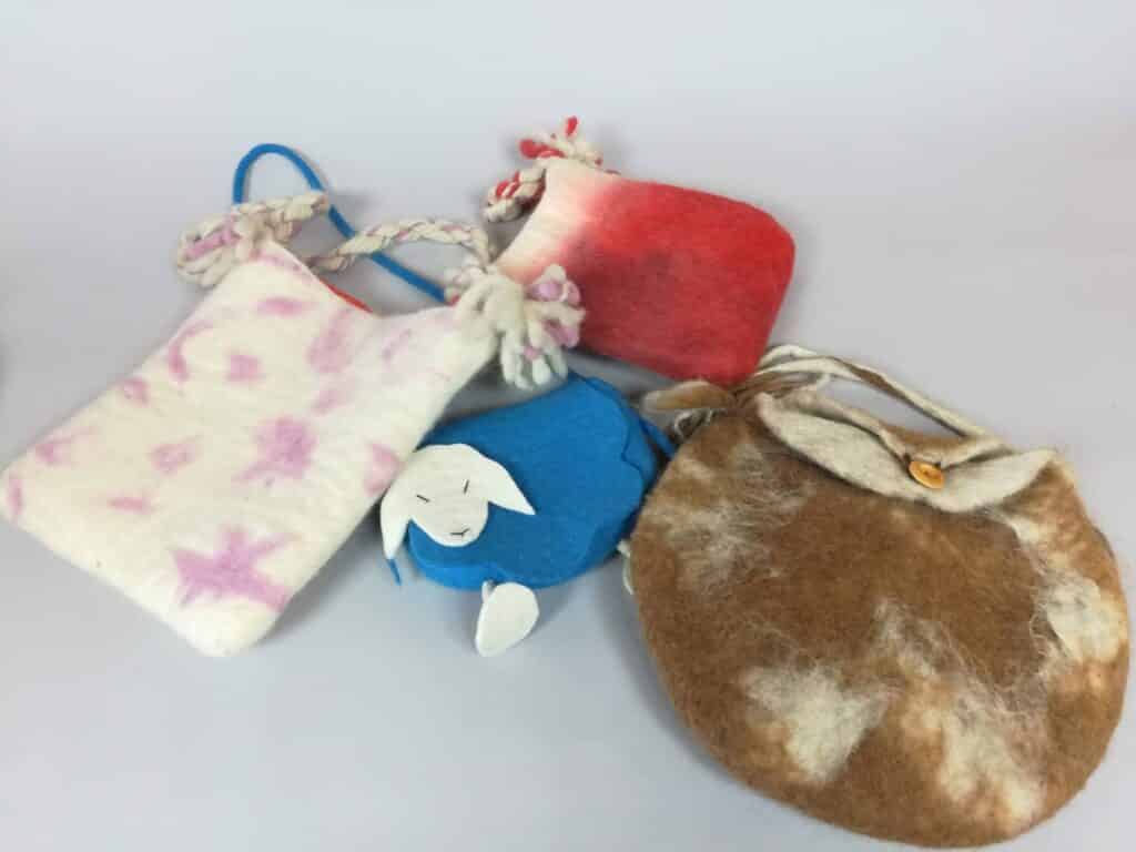Vier handgefertigte Filztaschen in verschiedenen Farben und Designs liegen auf einer grauen Oberfläche. Eine ist weiß mit rosa Sternen, eine rote mit weißen Enden, eine blaue mit einem Tierdesign und eine braun-weiße mit einem Knopfverschluss.