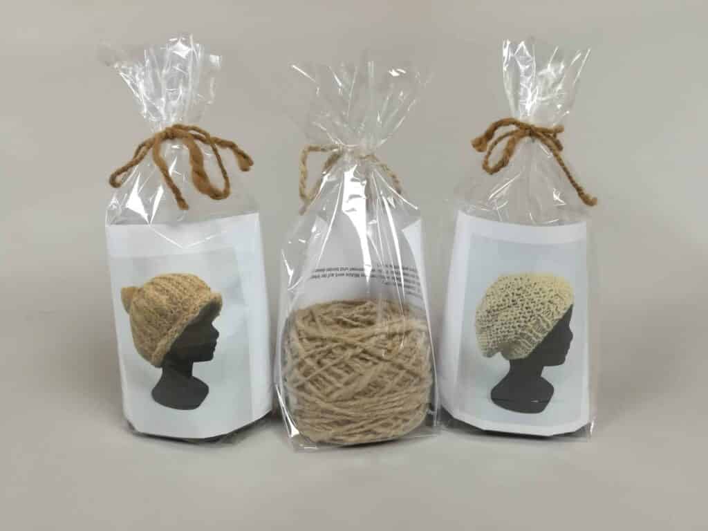 Drei kits in durchsichtigen Tüten mit Hanfschnur gebunden. Links und rechts Bilder von gehäkelten Mützen auf Schaumstoffköpfen, mittig ein Knäuel beiger Wolle.