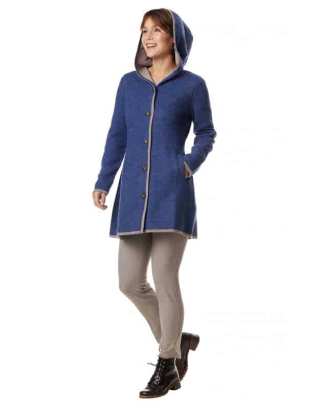 Eine lächelnde Person trägt einen blauen Kapuzenmantel mit Knöpfen über einer hellen Hose und schwarzen Stiefeletten.