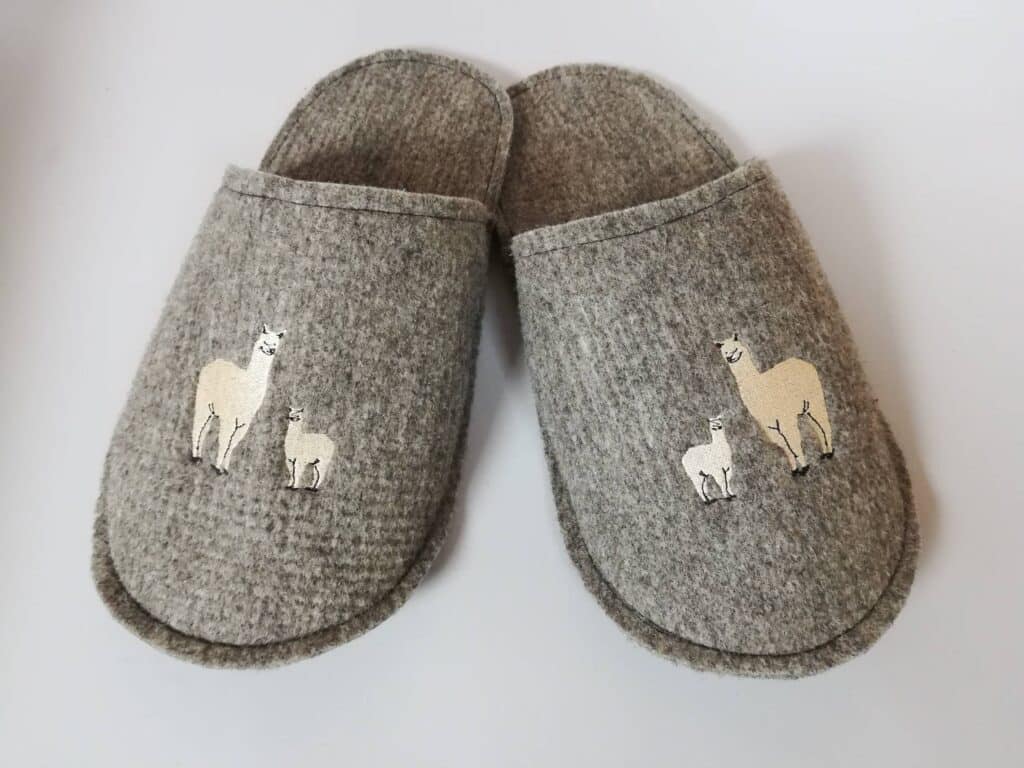 Ein Paar graue Filzpantoffeln mit aufgestickten Alpaka-Motiven, bestehend aus einer großen und einer kleinen Alpakafigur auf jedem Pantoffel.