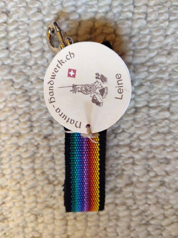 Farbiges Schlüsselband mit Regenbogenstreifen und einem runden Etikett, auf dem "natura-handwerk.ch" und "Leine" steht, auf einem hellen Teppich liegend.