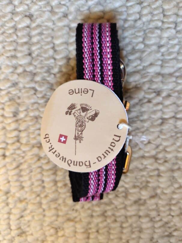 Ein schwarz-pink gestreiftes Stoffband mit einem runden, beigefarbenen Etikett, auf dem "Natur-Handwerk.ch" und "Leine" sowie das Symbol einer alten Spinnmaschine und eine kleine Schweizer Flagge abgebildet sind, liegt auf einem beigen Teppich.