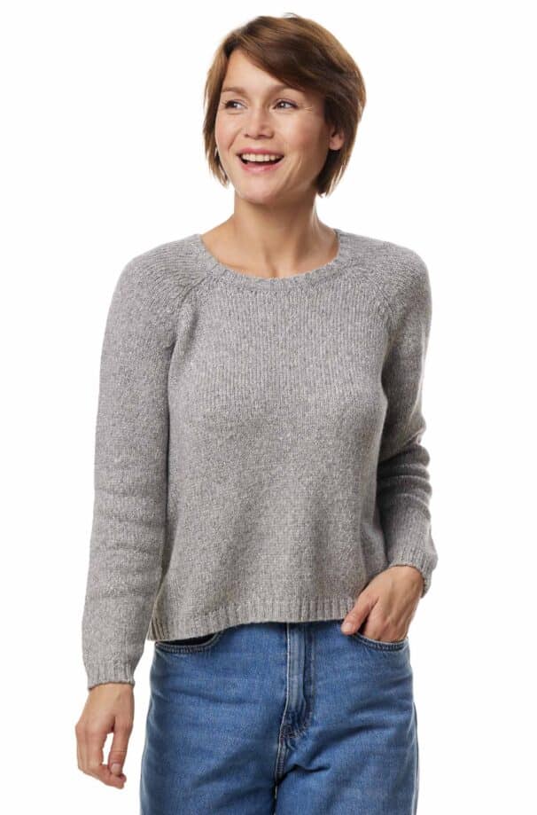 Eine lächelnde Person trägt einen grauen Pullover und blaue Jeans.