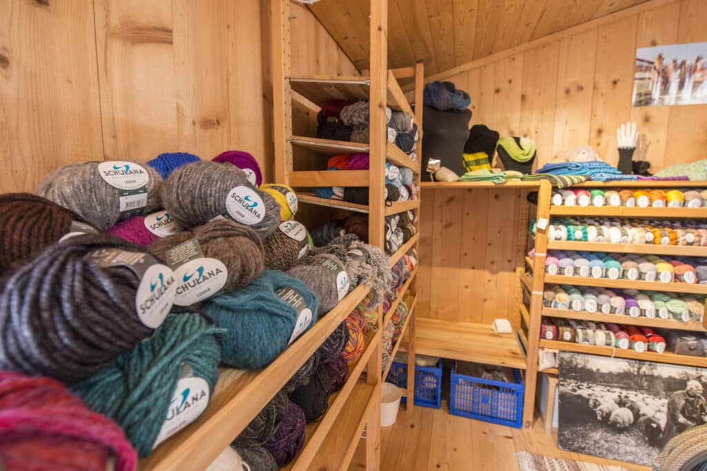 Ein kleiner Laden mit Regalen voller Wollknäuel in verschiedenen Farben, die ordentlich angeordnet sind. Die Wände und das Mobiliar sind aus hellem Holz gefertigt. Auf den Regalen steht "Schulana" auf den Etiketten der Wolle.