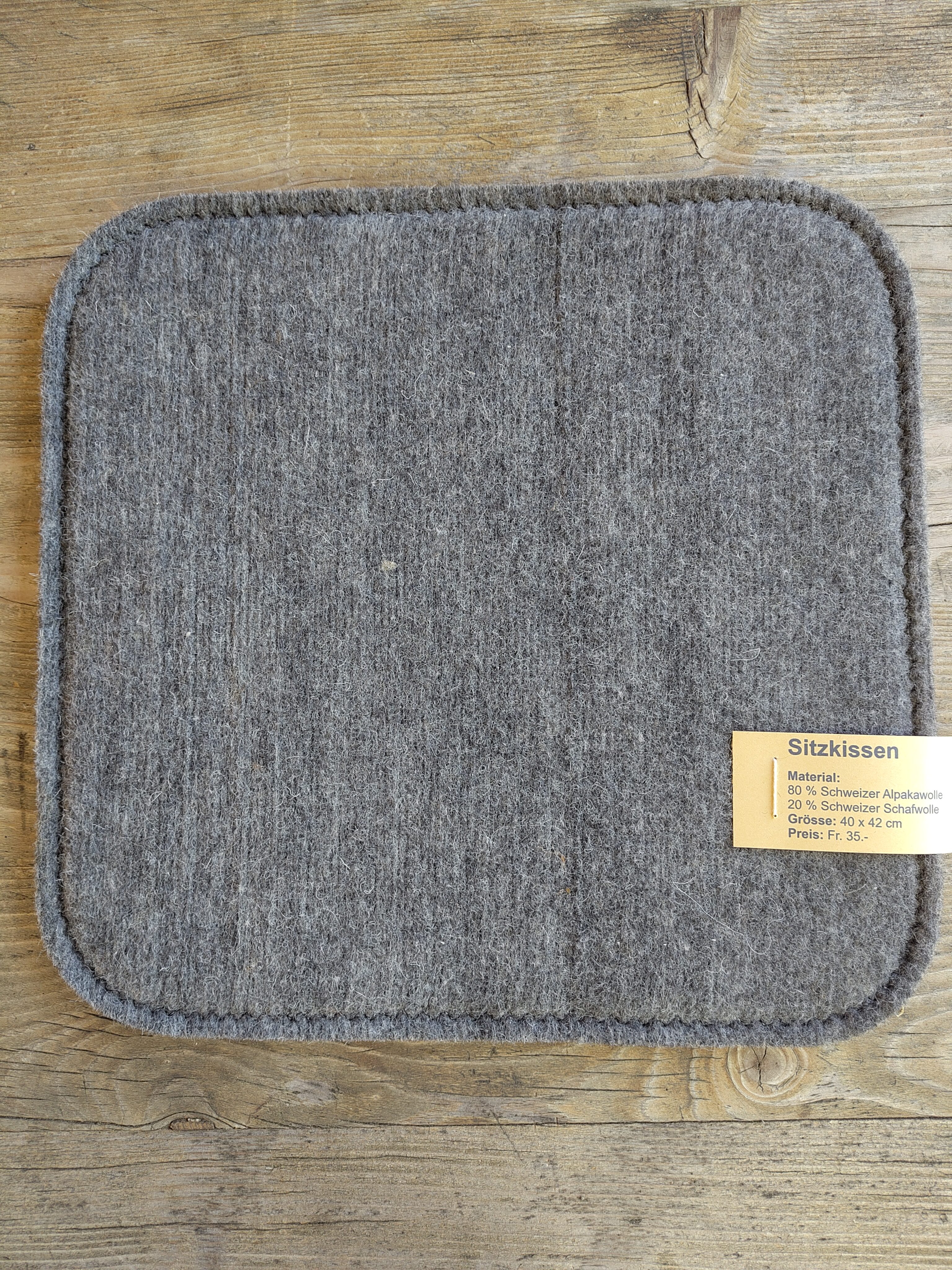 Graues Sitzkissen aus Wollfilz mit gelbem Etikett, das Material und Preis beschreibt, auf Holztisch liegend.