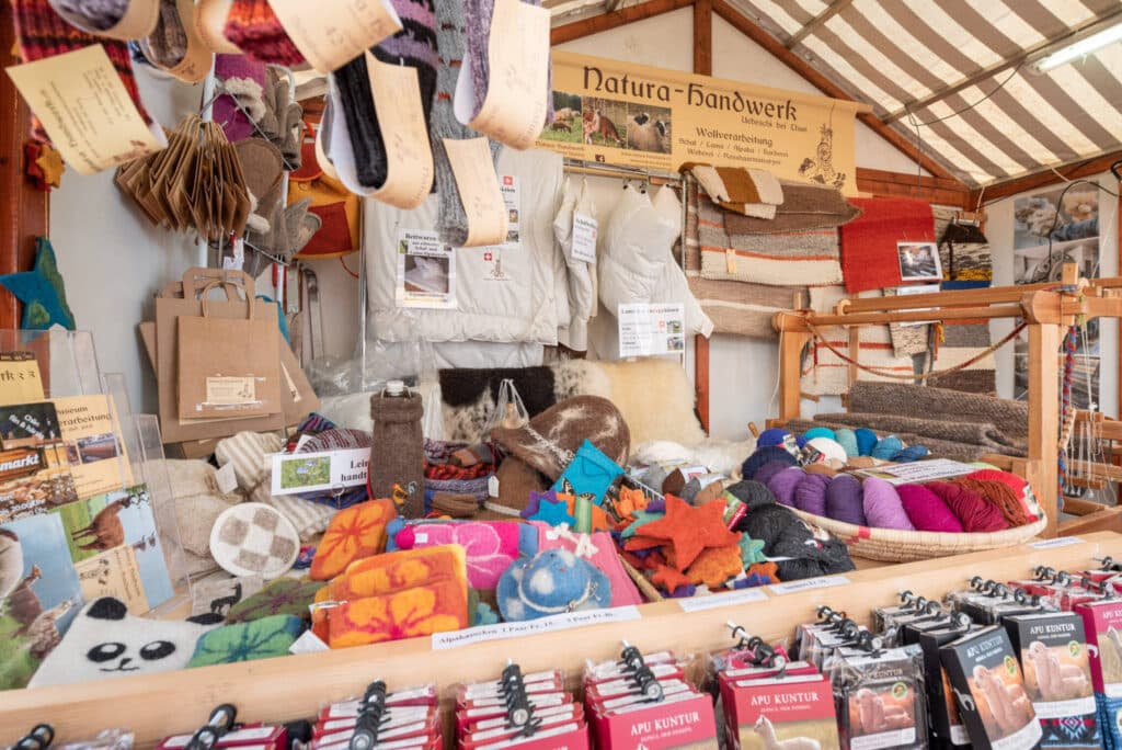 Marktstand mit handgefertigten Wollprodukten, darunter Socken, Schals, Pullover und Filzdekorationen, sowie ein Webstuhl im Hintergrund.