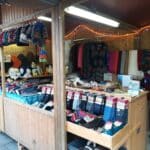 Verkaufsstand auf einem Markt mit Mützen, Pullovern, Socken und Schals; ein Verkäufer steht daneben.