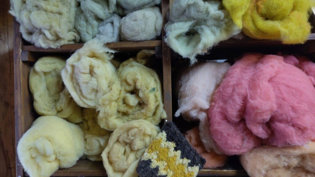 Farbenfrohe Kisten mit roher Wolle in verschiedenen Farbtönen, darunter Gelb, Grün, Rosa und Grau, sowie ein gestricktes Musterstück.