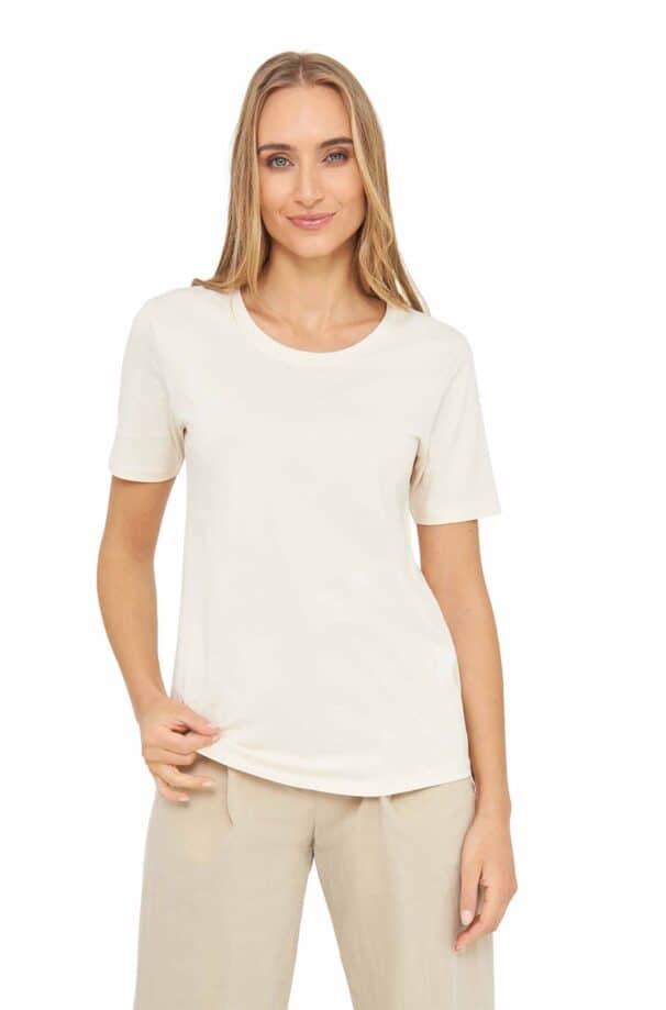 Frau in einem cremefarbenen T-Shirt und hellen Hosen vor weißem Hintergrund.