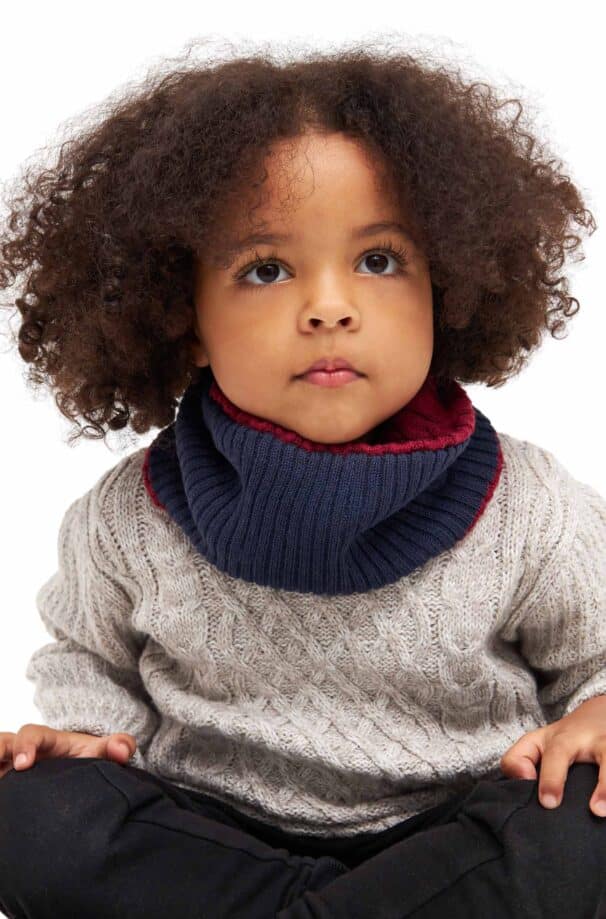 Ein Kind mit lockigem Haar trägt einen grauen Strickpullover und einen blauen Schal.
