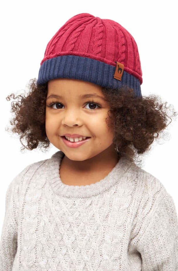 Ein Kind mit lockigem Haar trägt eine rot-blaue Strickmütze und einen hellgrauen Pullover mit Zopfmuster.