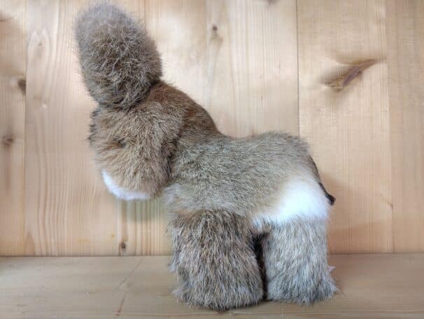 Ein Plüschtier, das wie ein Hase geformt ist, steht vor einer Holzwand. Es hat braunes und weißes Fell und steht aufrecht.