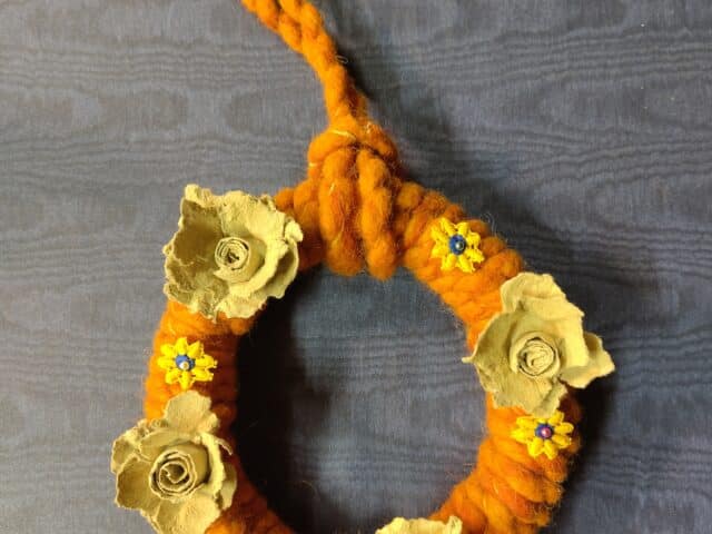 Orangefarbener, geflochtener Kranz mit beigefarbenen Papierblumen und kleinen blauen Blumen auf dunklem Hintergrund.