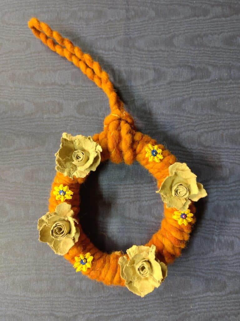 Orangefarbener, geflochtener Kranz mit beigefarbenen Papierblumen und kleinen blauen Blumen auf dunklem Hintergrund.