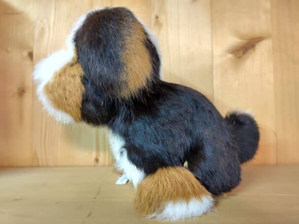 Ein dreifarbiges Plüschtier, das einen Hund darstellt, steht auf einem hölzernen Untergrund vor einer Holzwand.