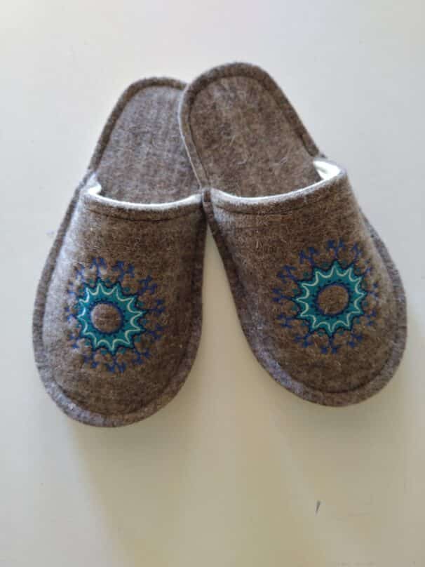 Ein Paar braune Filzpantoffeln mit blauer und grüner Stickerei in der Mitte, auf einer weißen Oberfläche liegend.