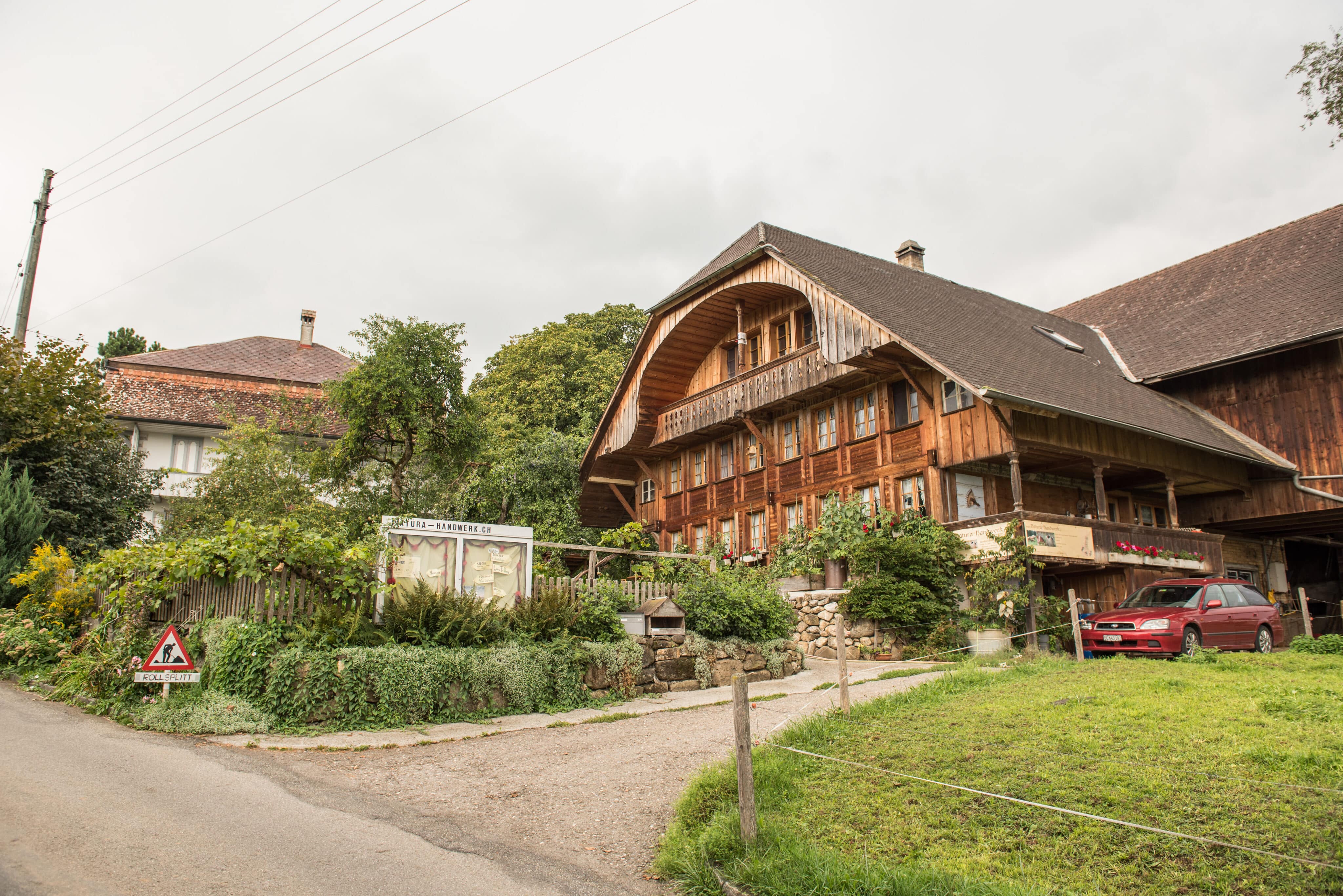 Ein traditionelles Schweizer Bauernhaus mit einem großen, hölzernen Satteldach und mehreren Fenstern. Vor dem Haus befindet sich ein Garten und ein rotes Auto ist rechts am Gebäude geparkt. Ein Schild mit der Aufschrift „Rollsplit“ befindet sich am Straßenrand, und in der Nähe steht ein weiteres Schild mit der URL „natura-handwerk.ch“.