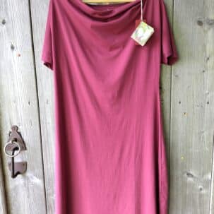 Ein bordeauxfarbenes Kleid hängt an einem Holzbügel vor einer rustikalen Holztür. Das Kleid hat kurze Ärmel und einen weiten Halsausschnitt. Ein Etikett mit blumigem Muster ist an der Kleidung befestigt.