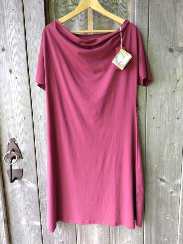 Ein bordeauxfarbenes Kleid hängt an einem Holzbügel vor einer rustikalen Holztür. Das Kleid hat kurze Ärmel und einen weiten Halsausschnitt. Ein Etikett mit blumigem Muster ist an der Kleidung befestigt.