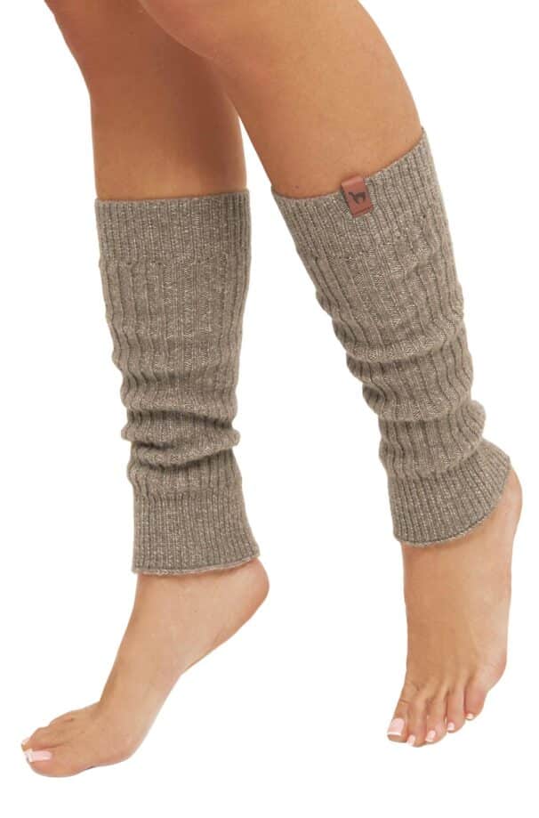 Ein Paar beige Strickstulpen an den Beinen einer Person, die auf Zehenspitzen steht, mit freiliegenden Füßen und einer kleinen braunen Marke oben auf der Stulpe.
