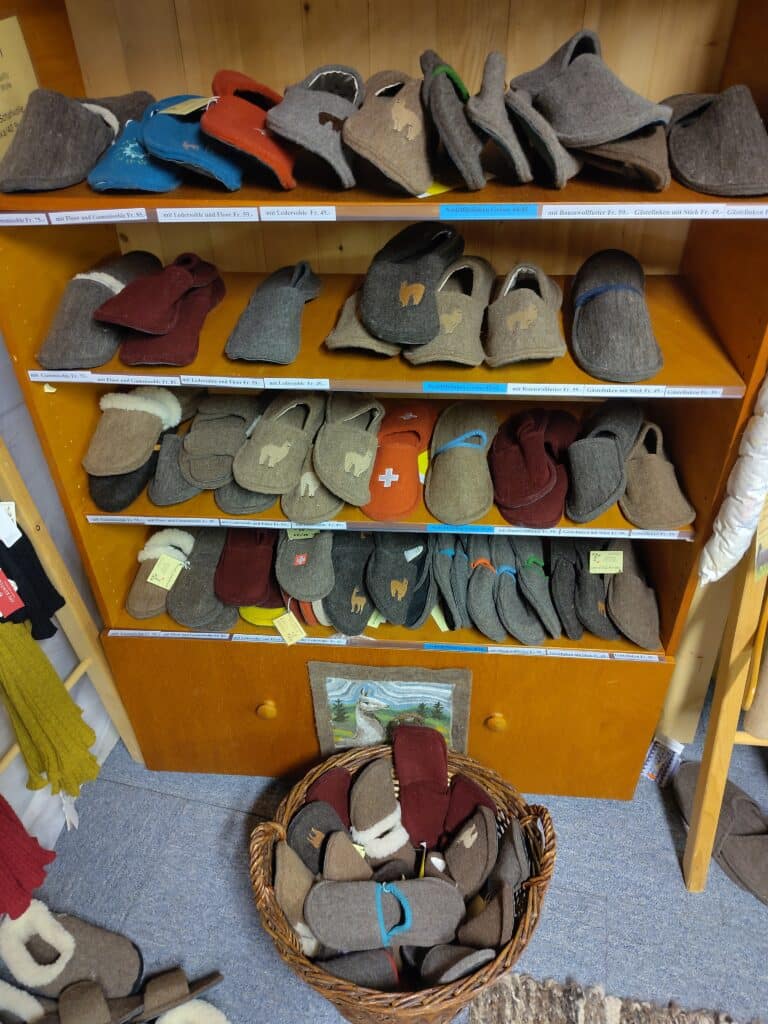 Ein Regal mit verschiedenen Paaren Filzpantoffeln in natürlichen Farbtönen, einige mit dekorativen Stickereien, darüber Preisschilder und darunter ein Korb mit mehr Pantoffeln.
