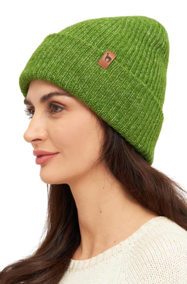 Frau trägt eine grüne Strickmütze mit einem kleinen braunen Logo auf der Seite, dazu einen cremefarbenen Pullover.