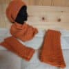 Ein Set aus orangefarbenem Strick, bestehend aus Mütze, Schal und Handschuhen, präsentiert auf einer schwarzen Schaufensterpuppe vor einem Holzpaneel-Hintergrund. Die Artikel haben ein Zopfmuster und ein Logo auf der Mütze.