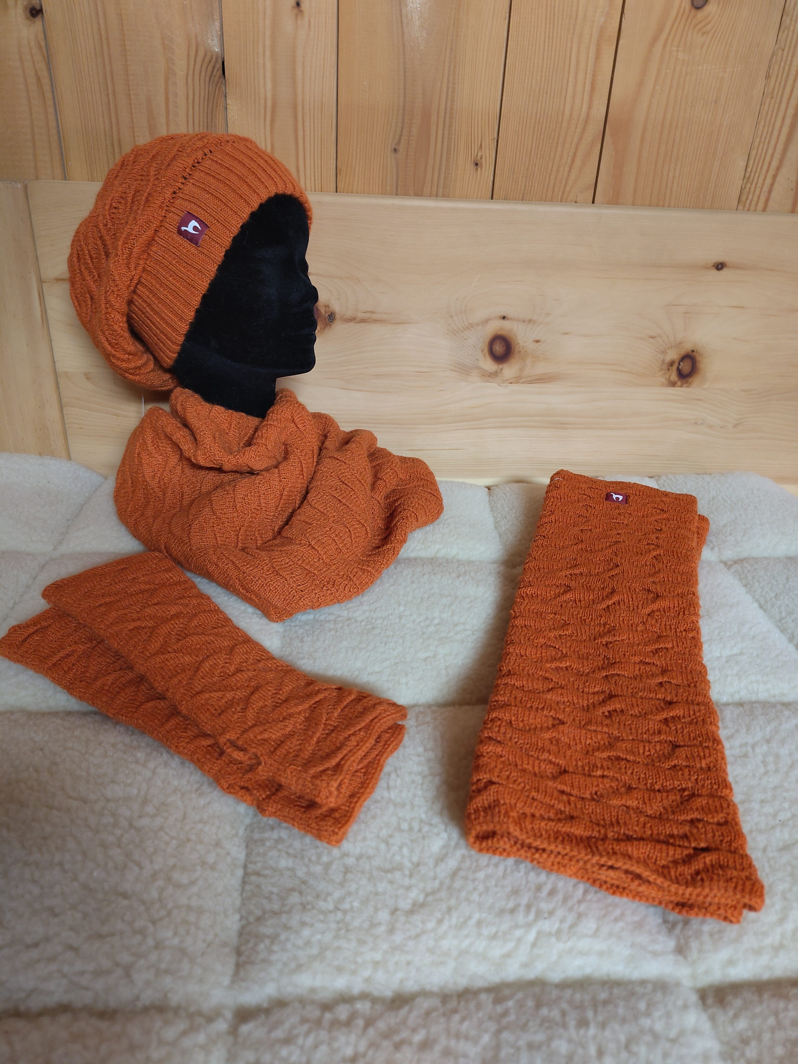 Ein Set aus orangefarbenem Strick, bestehend aus Mütze, Schal und Handschuhen, präsentiert auf einer schwarzen Schaufensterpuppe vor einem Holzpaneel-Hintergrund. Die Artikel haben ein Zopfmuster und ein Logo auf der Mütze.