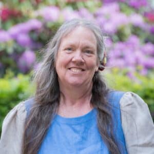 Eine lächelnde Frau mit langen grauen Haaren und einem blauen Kleid steht vor einem bunten Hintergrund aus verschwommenen Blumen.