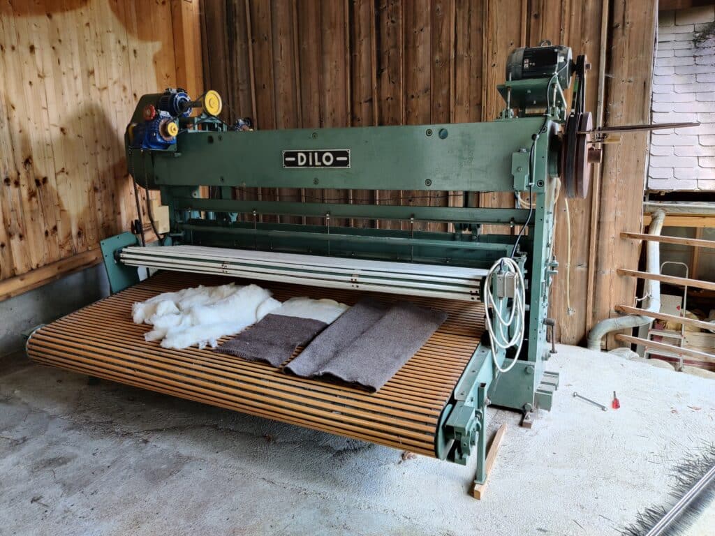 Eine grüne Industriemaschine von DILO in einem Holzraum, auf der weiße und braune Stoffbahnen liegen, mit gelben Bedienknöpfen und elektrischen Kabeln rechts.