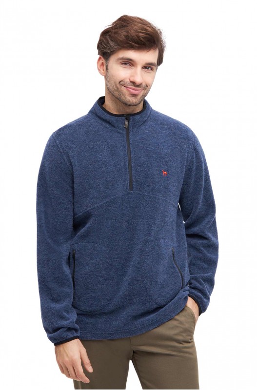 Ein Mann trägt einen blauen, langärmeligen Fleecepullover mit Reißverschluss am Kragen und einer kleinen roten Stickerei auf der Brust, kombiniert mit braunen Hosen.