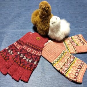 Zwei gestrickte Handschuhe mit bunten Mustern liegen neben zwei Plüsch-Alpakas auf einem blauen Hintergrund.