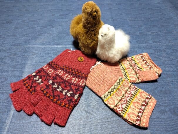Zwei gestrickte Handschuhe mit bunten Mustern liegen neben zwei Plüsch-Alpakas auf einem blauen Hintergrund.