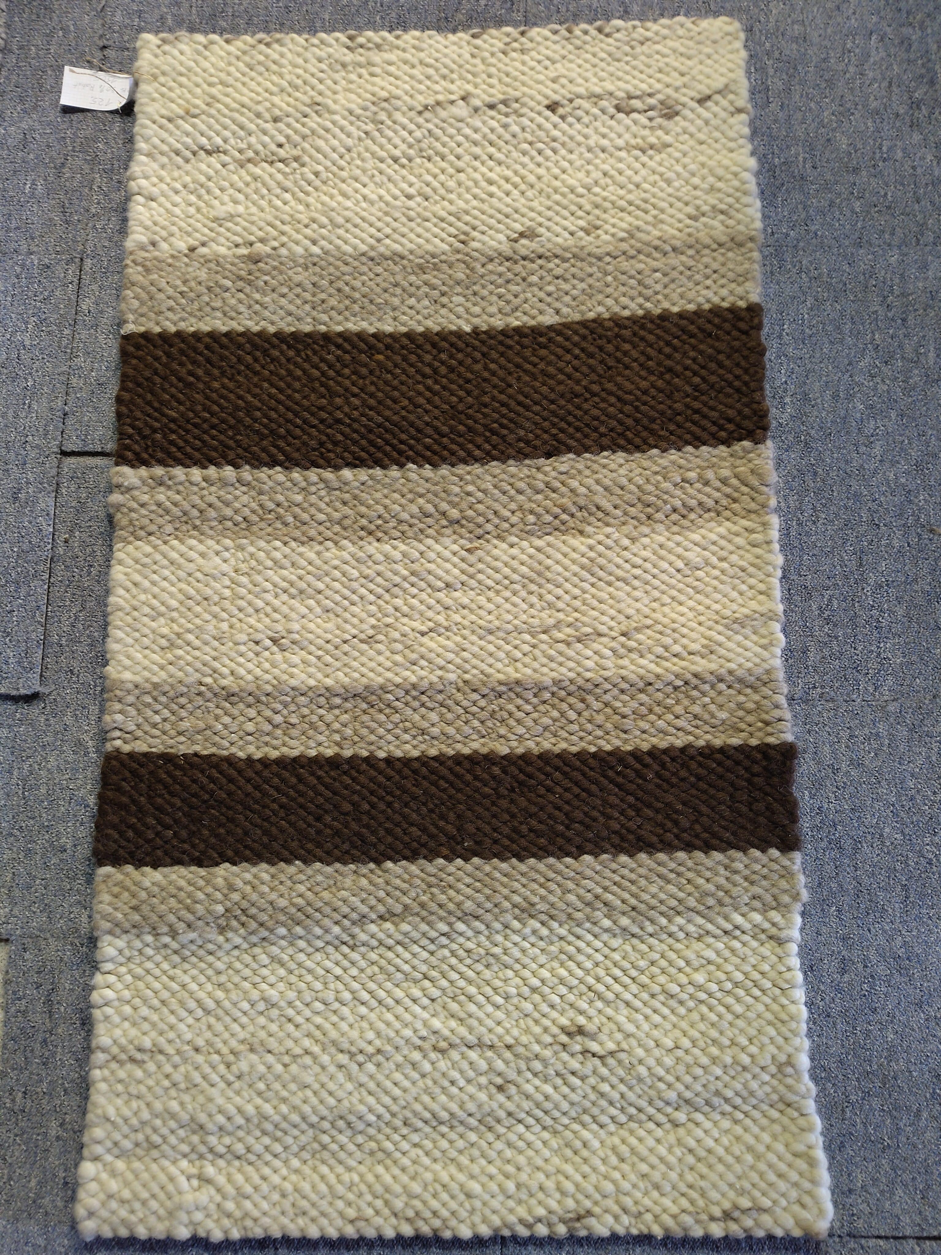 Handgewebter Teppich mit abwechselnden Streifen in Creme- und Brauntönen auf grauem Boden.