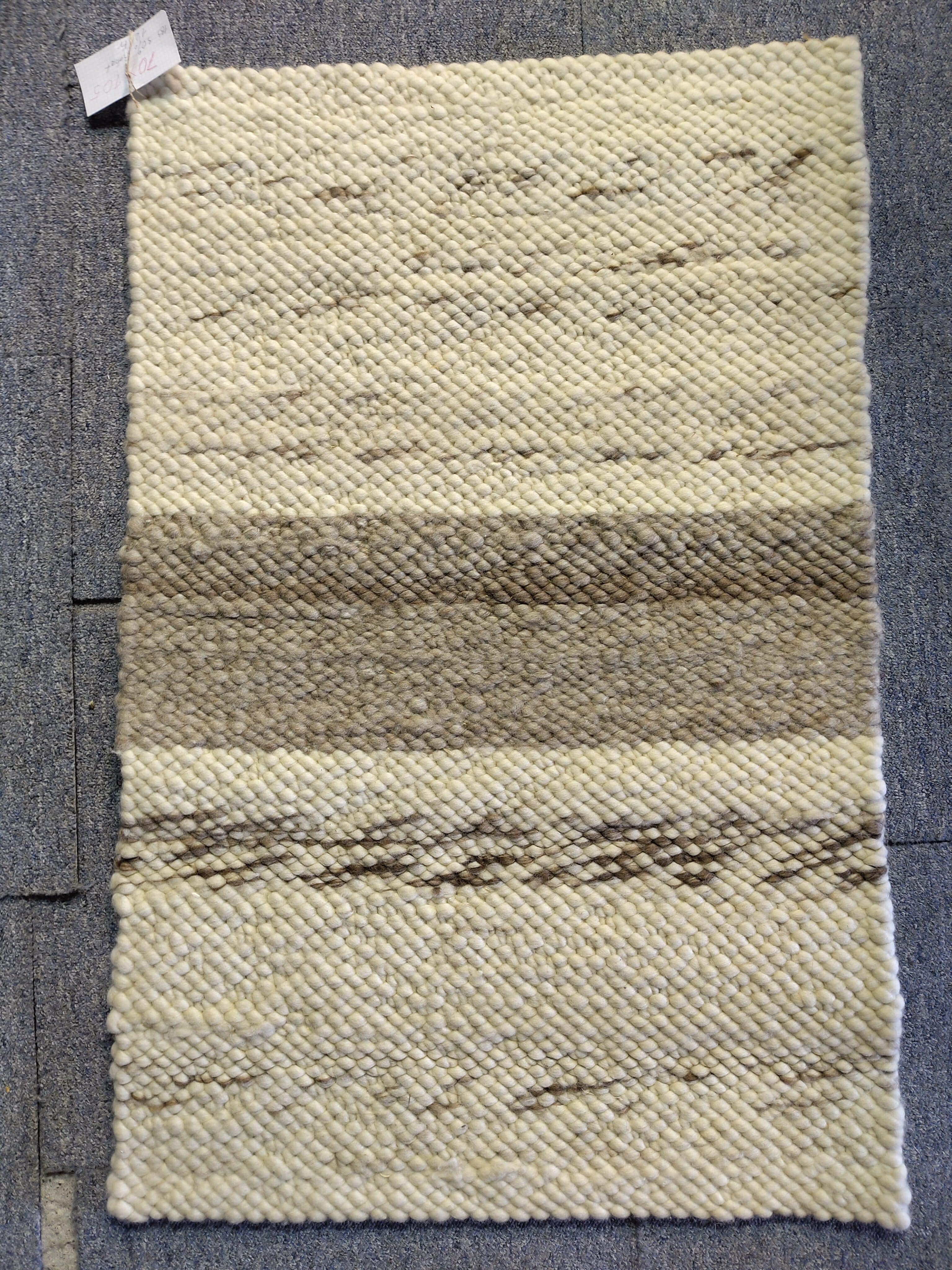 Ein handgewebter Teppich mit cremefarbenen und braunen Streifen, auf einem grauen Boden liegend.