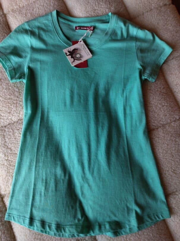 Grünes T-Shirt liegt flach auf einem beigen Stoff, mit Etikett am Kragen.