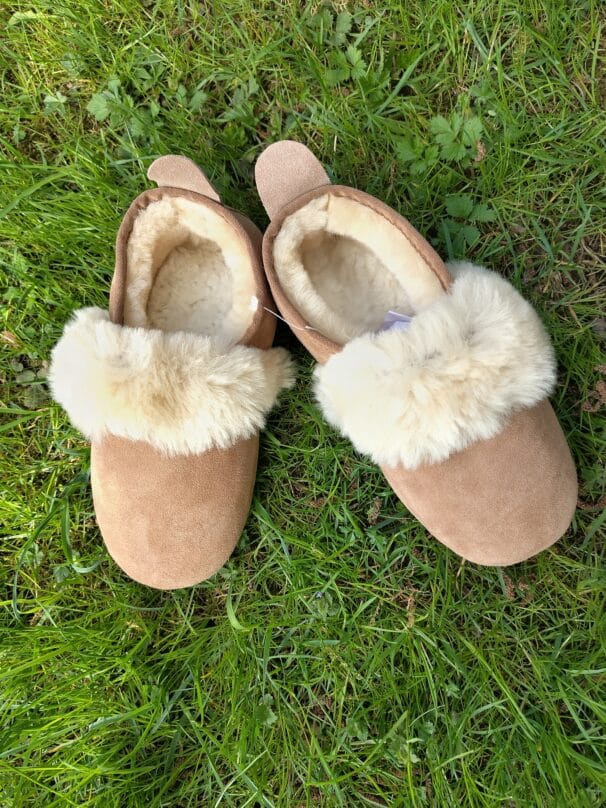 Ein Paar braune Hausschuhe mit flauschigem weißen Fellbesatz liegt auf grünem Gras.