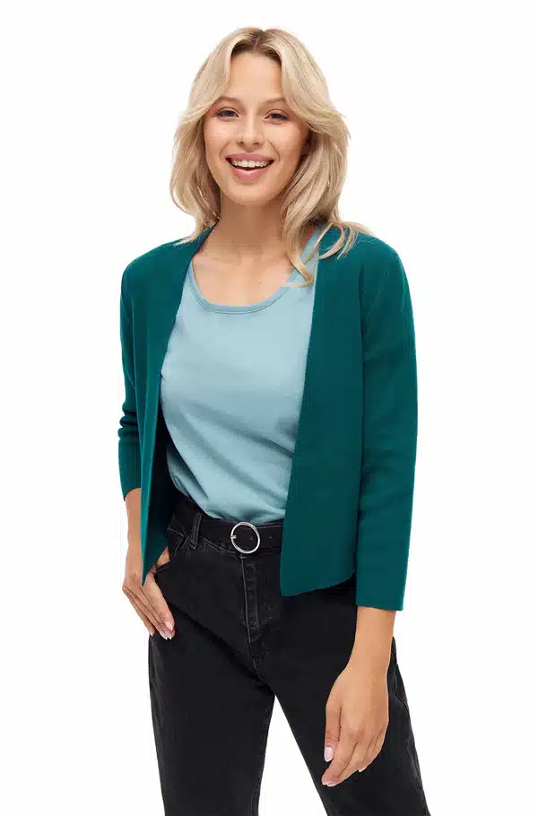 Eine lächelnde Frau in einem türkisfarbenen Oberteil und dunkelgrüner Strickjacke posiert freundlich.