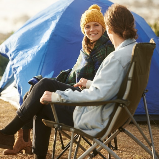 Zwei Personen sitzen vor einem blauen Zelt in einem Campingstuhl, eine trägt eine gelbe Mütze und lächelt.