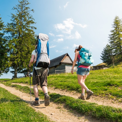Zwei Personen mit Rucksäcken wandern auf einem Pfad in einer ländlichen Landschaft mit Bäumen und einer Holzhütte im Hintergrund.