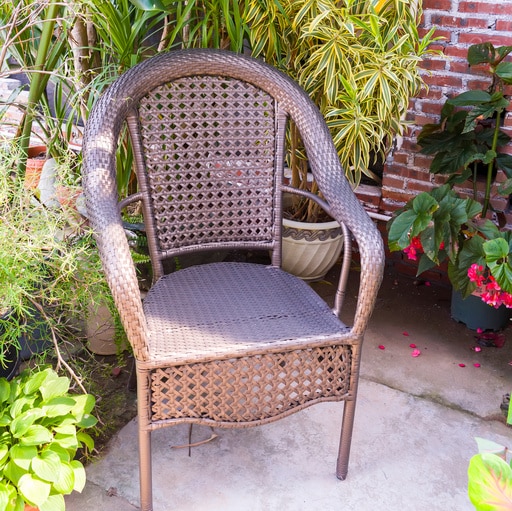 Ein brauner Korbstuhl steht auf einer Terrasse, umgeben von üppigem Grün und Blumentöpfen.