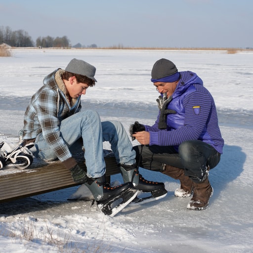 Zwei Personen ziehen auf einer schneebedeckten Holzplattform Schlittschuhe an, umgeben von einem gefrorenen See und winterlicher Landschaft.